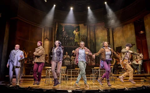 Six actors stand in a row on stage, holding hands and dancing, with musicians and two actors in the background. The set features wooden chairs, warm lighting, and a two-level rustic backdrop.