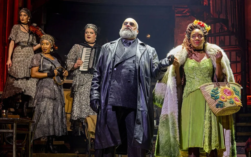 Five actors perform onstage in elaborate costumes; a man in a leather coat and sunglasses stands centre, holding a woman's arm, while three women in 1920s-inspired dresses and one with an accordion stand behind them.