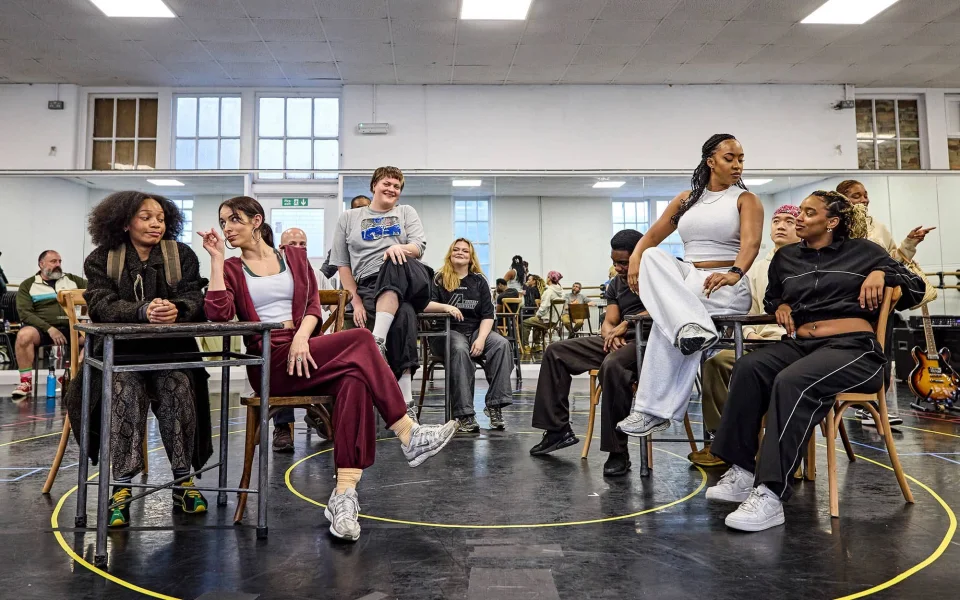A diverse group of performers in casual clothes sit and interact on chairs arranged in a rehearsal studio, with mirrors and musicians in the background. Some people are laughing and engaged in conversation.