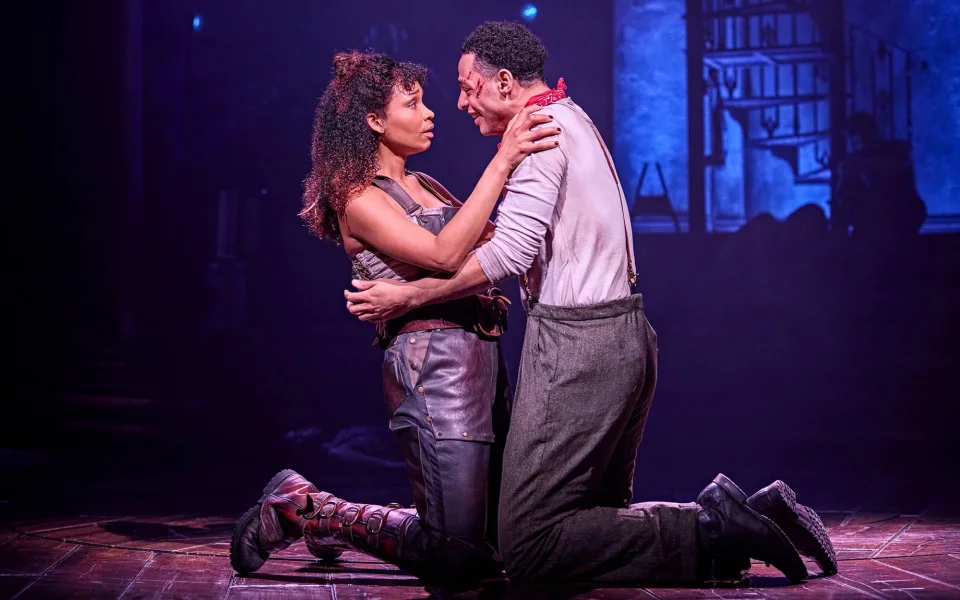 Two actors kneel on stage, holding each other closely and gazing into each other's eyes, under dramatic lighting with a blue-purple backdrop, suggesting an emotional or intense moment in a theatrical performance.