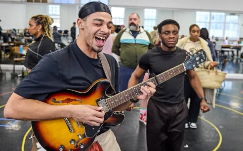A man joyfully plays an electric guitar in a lively rehearsal studio, surrounded by people talking and preparing in the background.