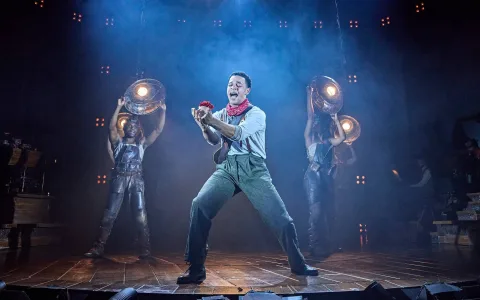 A stage performer in a red neckerchief passionately sings while holding a red flower, with two dancers in armor-like costumes and spotlights behind him, all set in dramatic blue stage lighting.