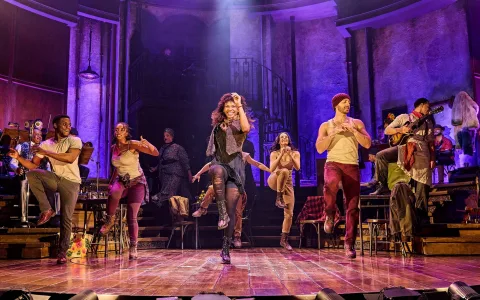 Actors perform a lively dance on stage with vibrant purple lighting; the central performer leads energetically while others surround her, dressed in urban costumes, with set pieces and stairs in the background.