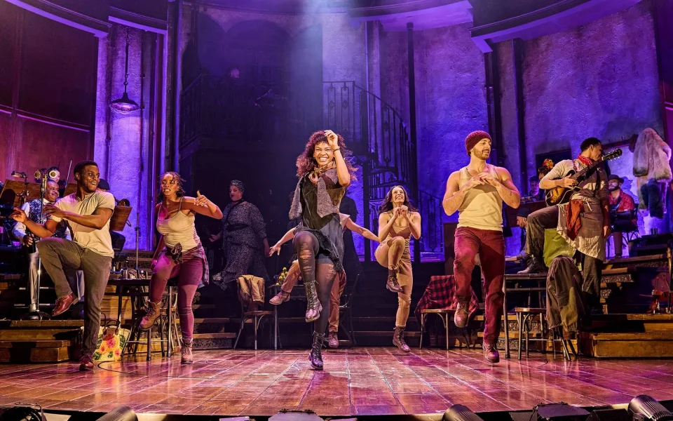 Actors perform a lively dance on stage with vibrant purple lighting; the central performer leads energetically while others surround her, dressed in urban costumes, with set pieces and stairs in the background.