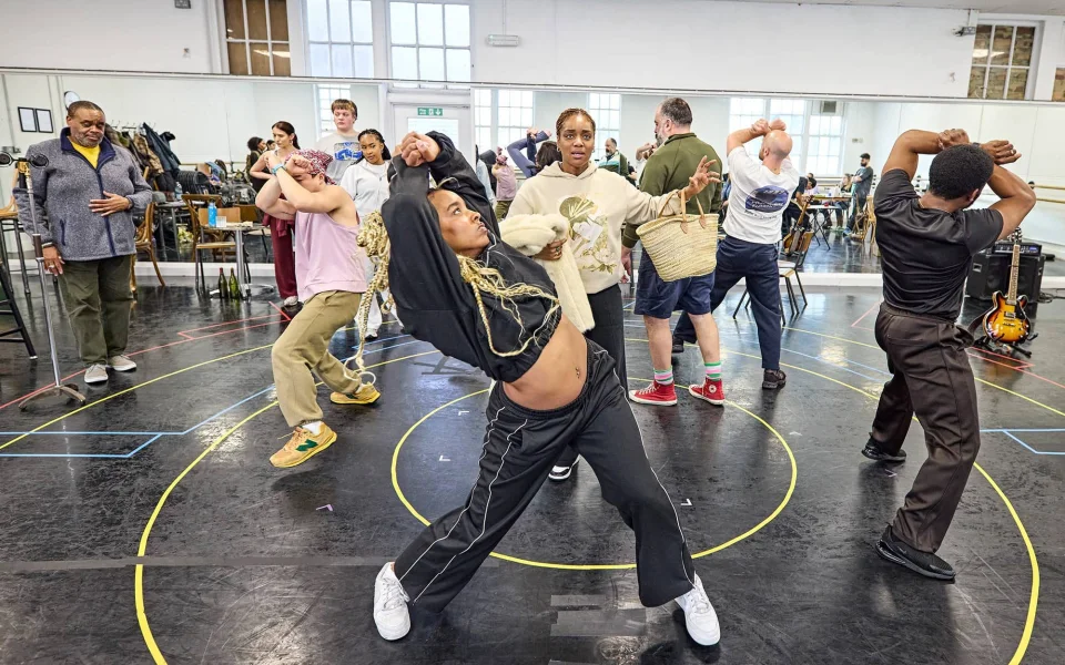 A group of people dance energetically in a rehearsal studio with mirrored walls. One person in the front stretches backward with arms out, while others pose and move around in various dance positions. Musical instruments are visible in the background.