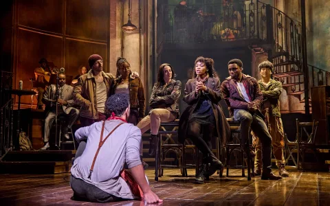 A group of actors on stage in a dramatic theater scene; one person sits on the floor facing seven others, who sit or stand close together, engaging in conversation under warm lighting and a detailed set backdrop.