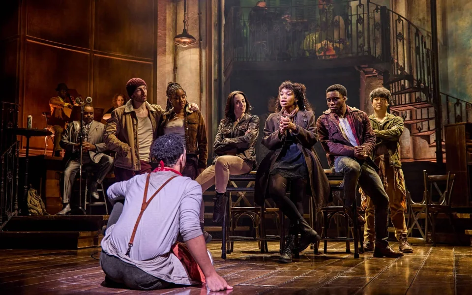 A group of actors on stage in a dramatic theater scene; one person sits on the floor facing seven others, who sit or stand close together, engaging in conversation under warm lighting and a detailed set backdrop.