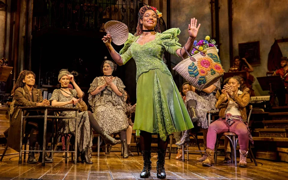 A performer in a green dress and boots stands center stage, smiling and holding a colorful basket, while other actors sit and clap in a lively theatrical setting with rustic décor.