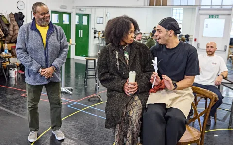 Three people interact in a rehearsal room. One man stands smiling, while a woman and another man sit together on chairs, engaged in conversation. A fourth person sits in the background, watching the scene.