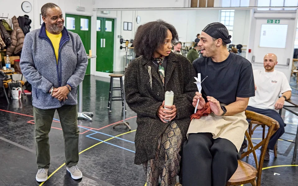 Three people interact in a rehearsal room. One man stands smiling, while a woman and another man sit together on chairs, engaged in conversation. A fourth person sits in the background, watching the scene.