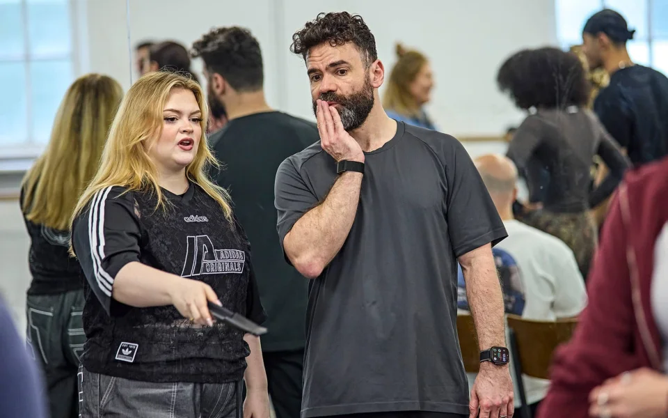 A woman gestures and speaks while holding a remote, as a man beside her listens with his hand on his chin. Several people are in the background of a bright, busy room.