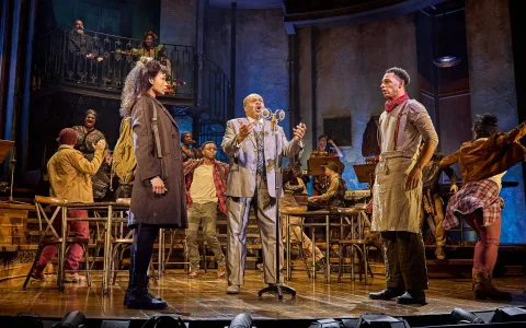 On a stage set resembling a rustic bar, a man in a suit sings at a vintage microphone while a woman in a coat and a man in an apron stand nearby; other cast members watch and react energetically in the background.