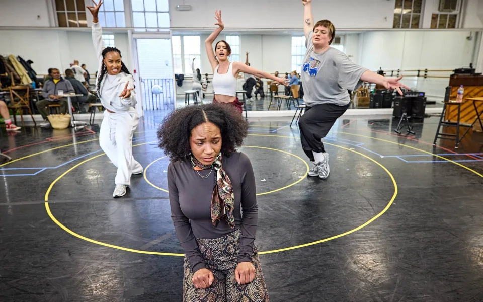 A woman sits on the floor looking upset while four people behind her dance energetically in a rehearsal studio with mirrors and musical instruments in the background.