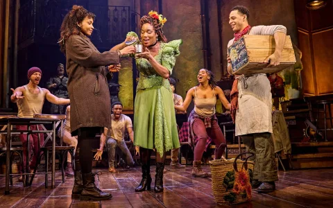 A lively stage scene from a musical: a woman in a green dress smiles as another woman in a coat offers her a small bouquet. Other cast members in colorful costumes react enthusiastically on a wooden set.