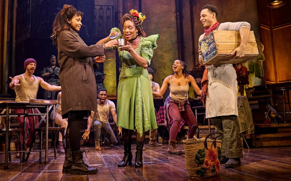 A lively stage scene from a musical: a woman in a green dress smiles as another woman in a coat offers her a small bouquet. Other cast members in colorful costumes react enthusiastically on a wooden set.