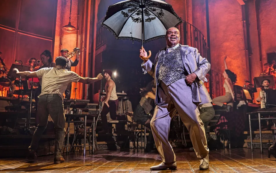 A joyful performer in a light purple suit holds a black-and-white parasol on stage, smiling, with musicians and dancers energetically performing in the background under warm stage lighting.