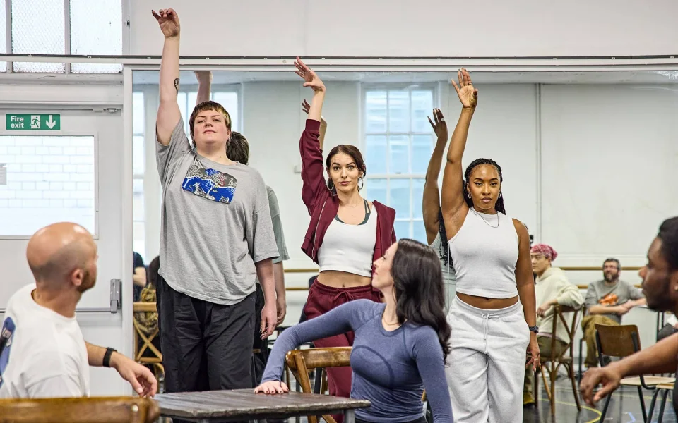 A group of people in casual clothing rehearse a dance routine in a studio, with three standing and raising one arm, while two others sit on chairs and watch or participate. A mirror reflects their movements in the background.