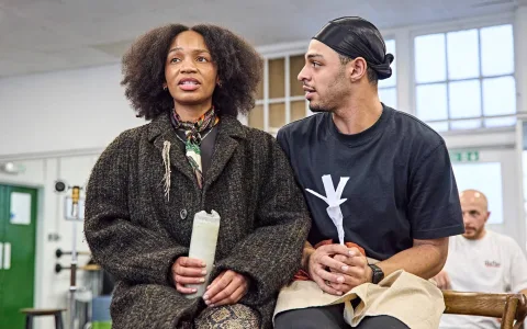 Two people sit close together indoors; the woman on the left wears a dark coat and holds a bottle, while the man on the right, in a black shirt and headscarf, looks at her and holds a white paper object. Another person is blurred in the background.