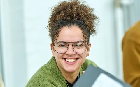 A person with curly hair, glasses, and a green sweater smiles. The background is blurred, focusing on their happy expression.