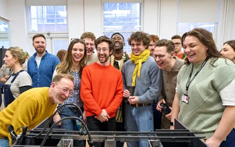 A group of people stand closely together in a bright room, smiling and laughing as they look at something out of view in front of them. Some are wearing colorful sweaters and glasses, and the atmosphere appears joyful and lively.