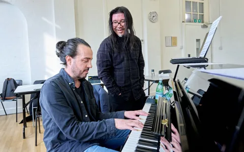 Two men in a bright room; one is seated playing the piano while the other stands nearby, smiling and watching. Sheet music and a bottle of water are on the piano. Tables and chairs are in the background.