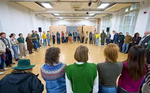 A large group of people stand in a spacious, brightly lit room, forming a wide circle. They are in rehearsal for a new musical, with some smiling and looking towards the center.