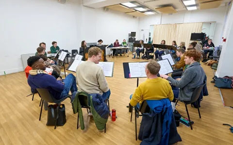 A group of people sit in a circle in a bright rehearsal room, each with music stands and sheet music. Some play instruments, others observe or take notes. The room has wooden floors and white walls.