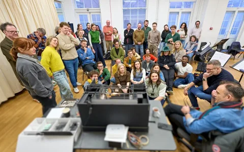 A large group of people are gathered in a bright room, standing and sitting around a table, listening attentively to a person talking through the stage design model box. Most are smiling or engaged.