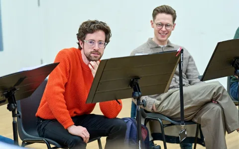 Two men sit in chairs behind music stands, listening attentively. One wears an orange sweater and glasses, leaning forward with his hand on his chin. The other, also in glasses, wears a beige sweater and smiles.