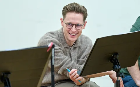 A smiling person with short, neatly styled hair and glasses sits holding a pen, looking toward someone off-camera. They are wearing a beige sweater and are seated in front of a music stand with sheets of paper.