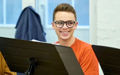 A young person wearing glasses and an orange shirt smiles while sitting behind a music stand in a bright room with a coat hanging in the background.