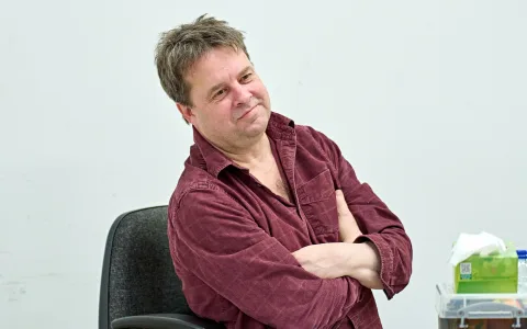A man with short brown hair wearing a maroon button-up shirt sits on a chair with his arms crossed, smiling slightly. The background is plain and white, with a tissue box and some papers on a table nearby.