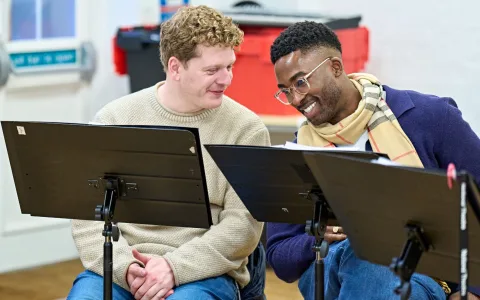 Two men sitting side by side smile and talk while looking at scripts on music stands in a rehearsal room. One wears a beige sweater; the other wears glasses, a scarf, and a blue cardigan.