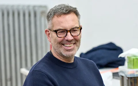 A man with short gray hair and a beard, wearing glasses and a navy blue sweater, smiles while sitting indoors. The background shows a radiator and some folded clothes.