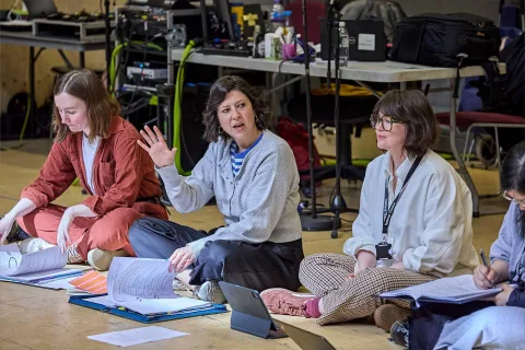 Three women sit on the floor with papers, laptops, and folders, engaged in discussion; equipment and cables are visible in the background.