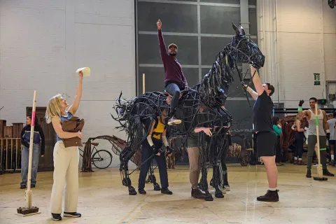 Actors rehearse with a large, black, horse puppet on stage; one person rides it as others operate and guide the puppet, whilst others observe or gesture nearby.