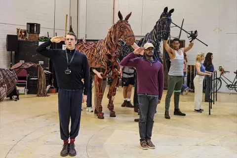 Actors rehearse with life-sized horse puppets; two men in front salute while others operate or stand near the puppets in a spacious rehearsal room.