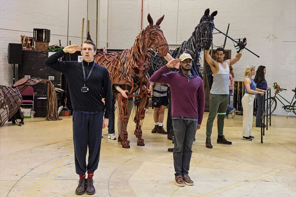 Actors rehearse with life-sized horse puppets; two men in front salute while others operate or stand near the puppets in a spacious rehearsal room.