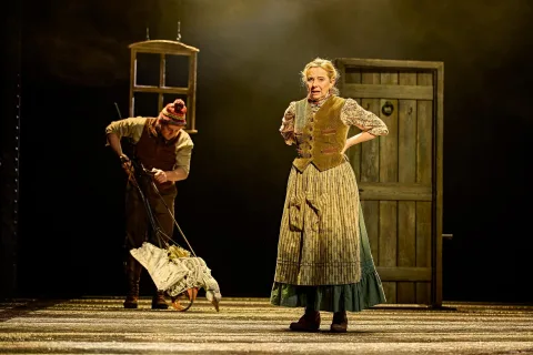 An older woman in period costume stands on stage, whilst an actor in the background walks a goose puppet on wheels. A wooden door and hanging window frame are visible.