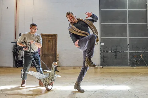 Two men rehearse, one pulls a prop swan on wheels while the other, wearing a waistcoat, lifts his leg dramatically as if reacting to the swan. A bicycle and door sit in the background.