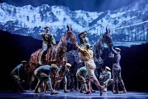 Actors on stage manipulate two large horse puppets, portraying soldiers and horses in a dramatic war scene with a battlefield backdrop. Two actors ride on top of the horses with swords drawn.
