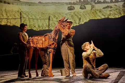 Three actors on stage control a horse puppet; one actor sits on the ground whistling using cupped hands. A rural landscape is projected in the background.
