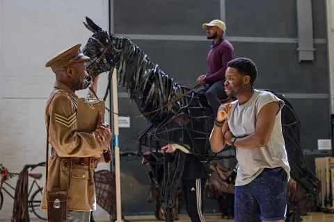 Two men talk in front of a large horse puppet, with another man sitting on the puppet. The setting appears to be a rehearsal space or backstage area.