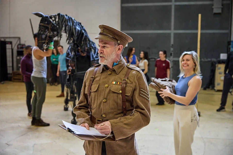 An actor in a soldier’s uniform holds a clipboard on stage, whilst others rehearse with a large horse puppet and props in the background.