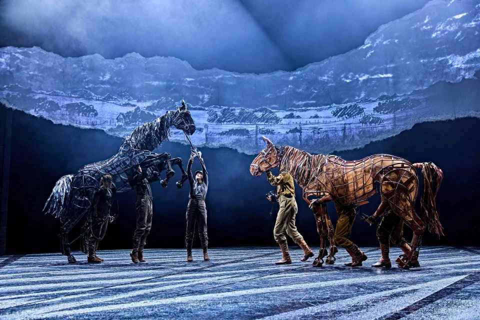 Actors operate two life-sized horse puppets on stage under dramatic blue lighting, with a backdrop depicting a scenic landscape.