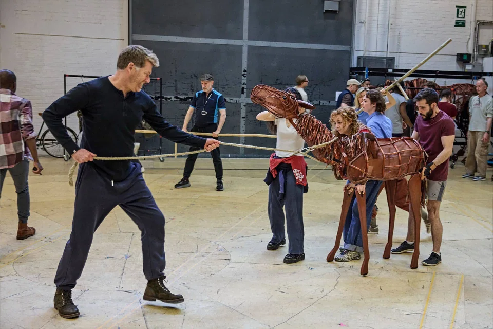 A group of people rehearses with a large horse puppet in a spacious studio, with one person pulling a rope in front.