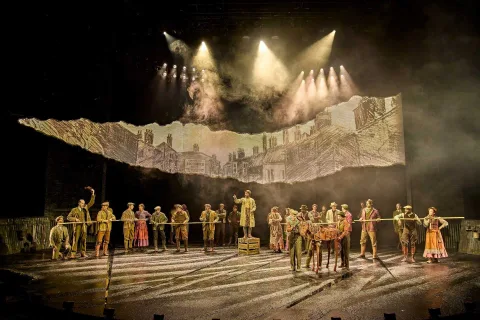 A large group of actors in period costumes stand on stage under dramatic lighting, with a torn backdrop depicting old city buildings behind them. Two puppeteers man a small horse puppet.