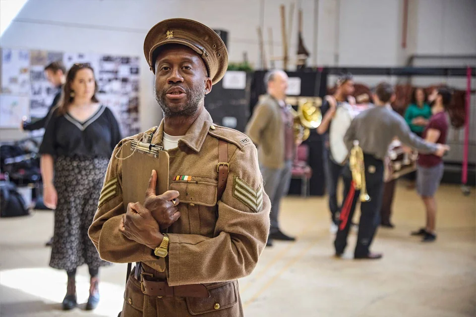 A man in a vintage military uniform stands in the foreground; behind him, people rehearse in a spacious room with musical instruments and scattered props.