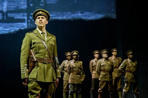 A group of men in historical military uniforms stand on stage, with one man in the foreground looking serious under dramatic lighting.