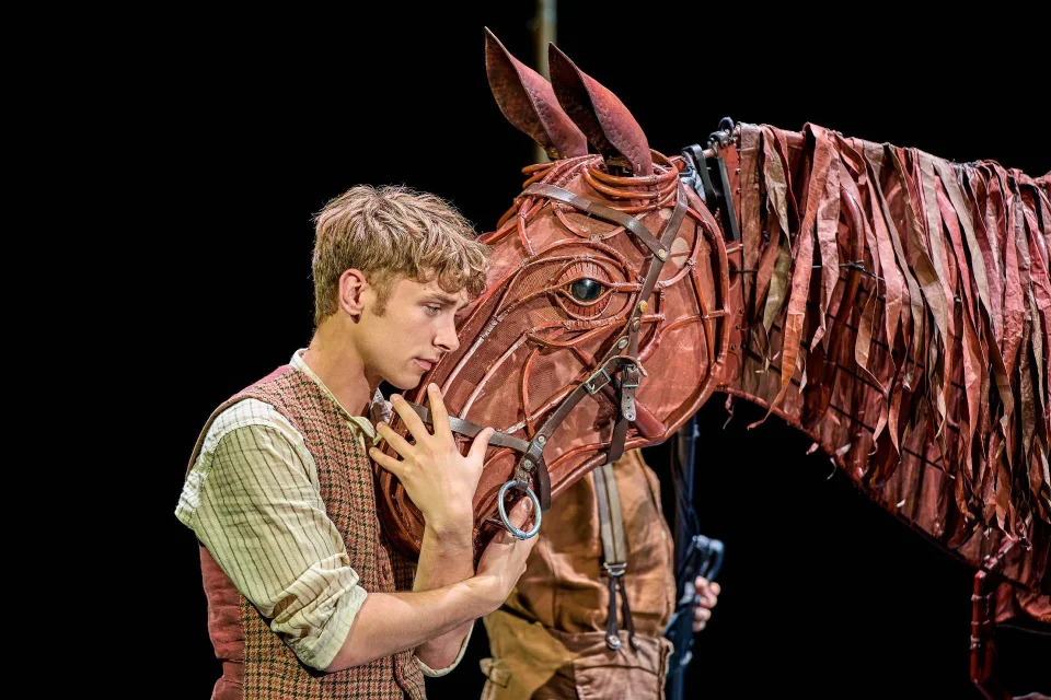 A young man gently touches the face of a life-sized horse puppet made of brown materials, with a puppeteer partly visible behind it.